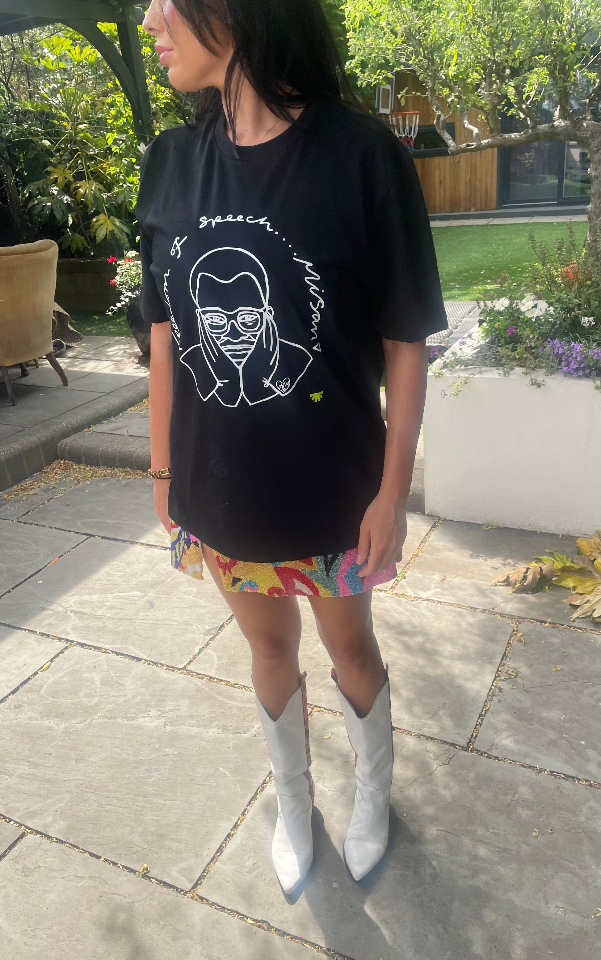 Person wearing a black t-shirt with a graphic design, colorful skirt, and white boots outdoors.